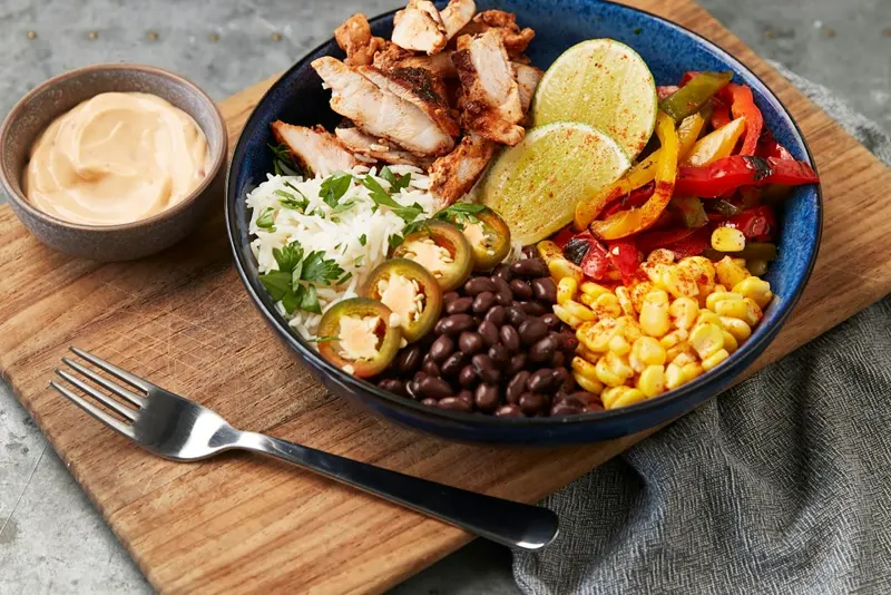 A burrito bowl with sliced chicken, rice, black beans, corn, peppers and lime
